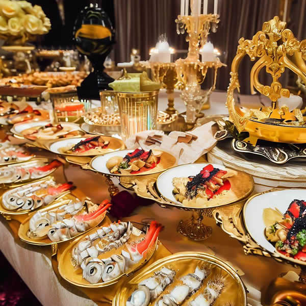 Un buffet d'assiettes en or et en porcelaine remplie de mets délicats, avec des bougies sur des socles dorés, un bouquet de rose jaune en fond.